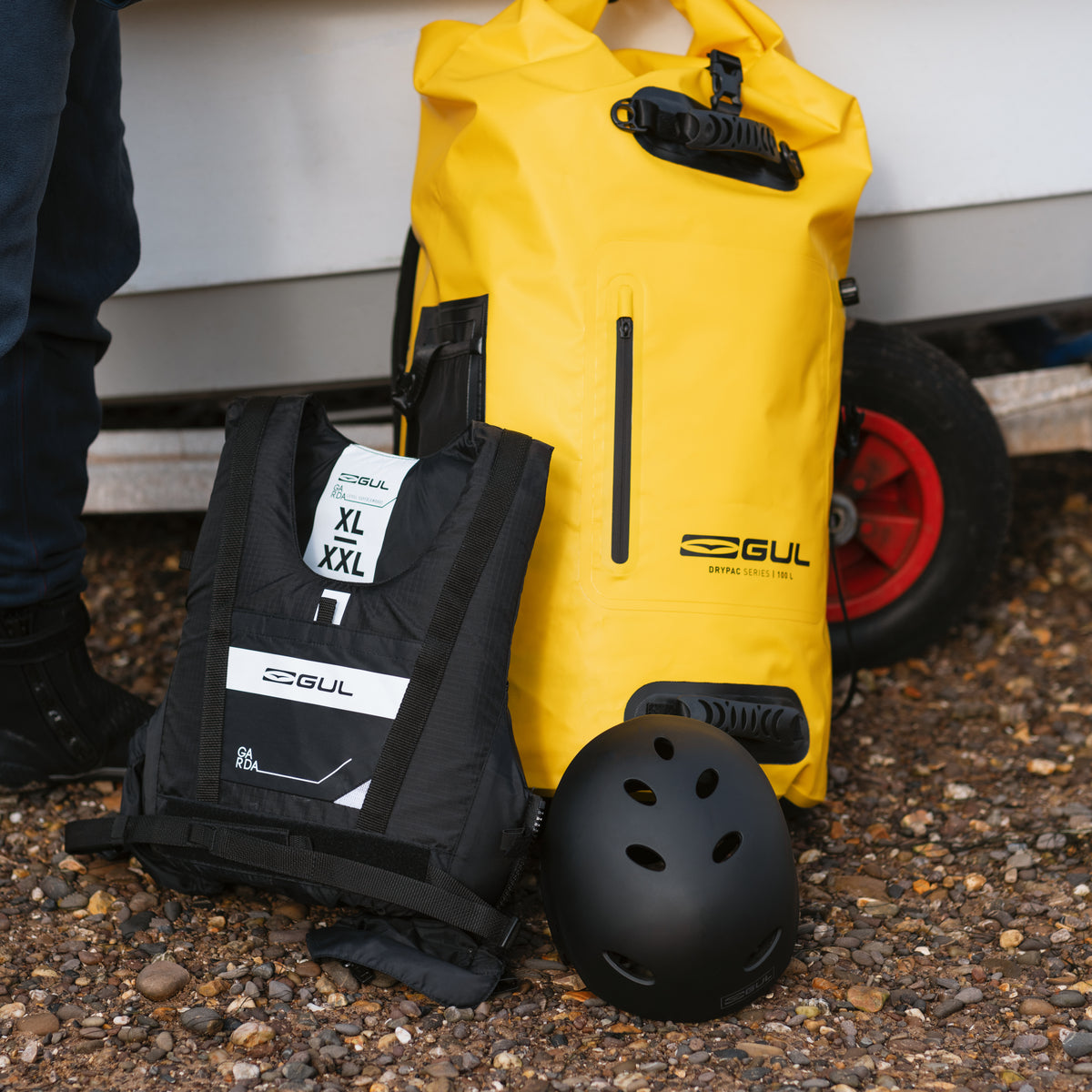 Yellow GUL Drypac 100L dry bag beside a buoyancy aid and helmet next to a boat.